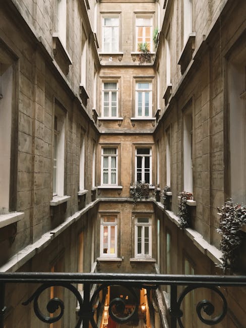 A narrow courtyard garden situated between tall, classical-style sandstone buildings with multiple white-framed sash windows. The courtyard features a lower level with a reflective pool or pond, bordered by a black wrought iron railing in the foreground. Along the edges of the upper level, small potted shrubs with dark purple leaves are placed on window sills and ledges. The overall scene is illuminated by natural daylight, highlighting the textures of the wet stone walls, trimmed shrubs, and the calm surface of the water, creating a serene and compact outdoor space suited for maintenance and landscaping work by gardeners Chelsea.